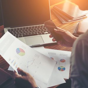 Businessman hands using cell phone with financial report graph at office desk.